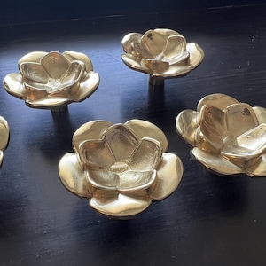 Może przedstawiać: Zestaw pięciu mosiężnych gałek do szafek w kształcie kwiatów. Każda gałka ma szczegółowy kwiatowy wzór z wieloma płatkami. Gałki są prezentowane na ciemnej powierzchni, podkreślając ich metaliczny połysk.