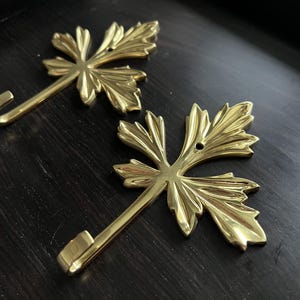 May include: Two gold leaf-shaped hooks with a decorative design. The hooks are attached to a dark surface.