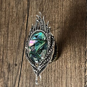 May include: A silver brooch featuring an oval abalone shell centre with iridescent green, blue, and purple hues. The brooch has an elaborate, leaf-shaped design with small, decorative details. The brooch is made of silver-toned metal.