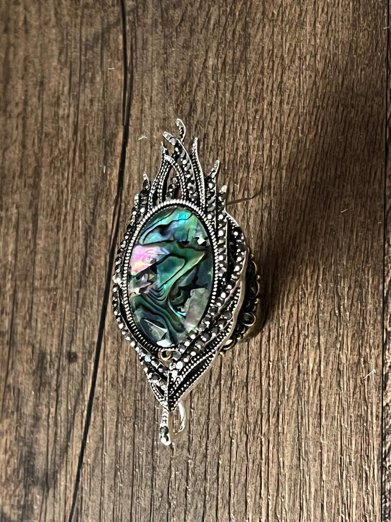 May include: A silver brooch featuring an oval abalone shell centre with iridescent green, blue, and purple hues. The brooch has an elaborate, leaf-shaped design with small, decorative details. The brooch is made of silver-toned metal.
