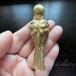 May include: A gold-coloured metal bird figurine with detailed feathers. The bird is facing away from the viewer, with its wings folded and its tail feathers spread out.