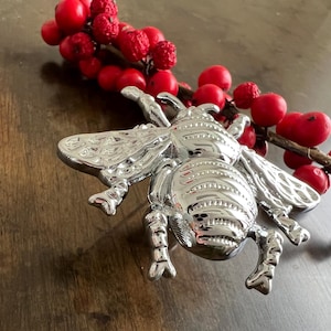 Könnte beinhalten: Ein silbernes Bienen-Ornament mit einem detaillierten Design. Die Biene sitzt auf einer Holzoberfläche mit einem Zweig roter Beeren.
