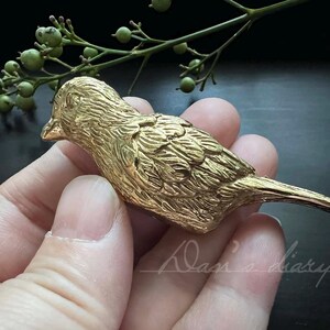 Puede incluir: Una figurita de p&aacute;jaro de metal de color dorado con marcas detalladas de plumas. El p&aacute;jaro est&aacute; posado en una mano, con las alas extendidas.