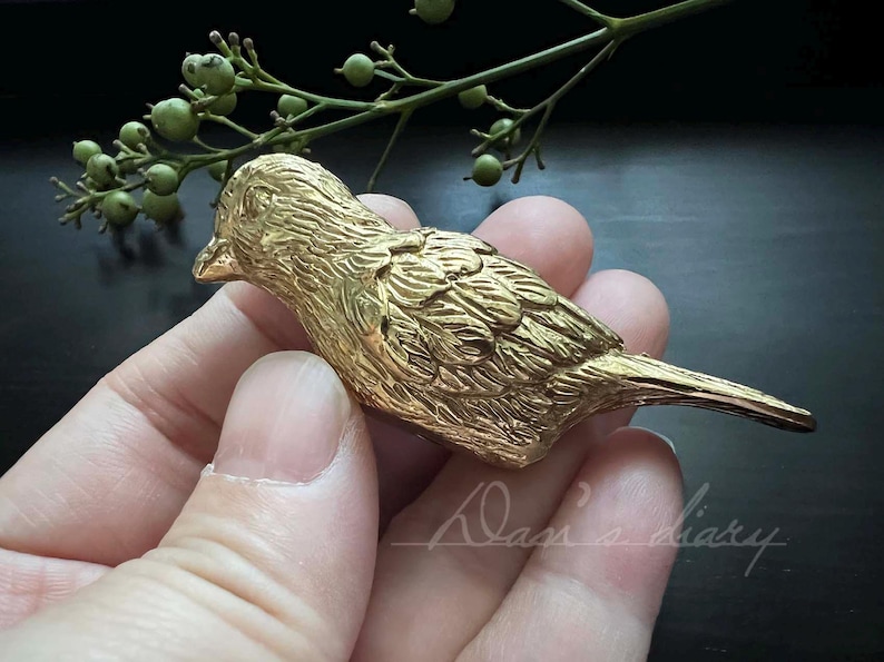 Puede incluir: Una figurita de p&aacute;jaro de metal de color dorado con marcas detalladas de plumas. El p&aacute;jaro est&aacute; posado en una mano, con las alas extendidas.