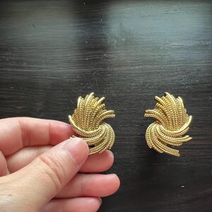 K&ouml;nnte beinhalten: Ein Paar goldfarbene Clip-Ohrringe. Jeder Ohrring hat ein strukturiertes, wirbelndes Design mit einer Ansammlung kleiner, vertikaler Linien. Die Ohrringe werden auf einer dunklen Holzoberfl&auml;che pr&auml;sentiert.