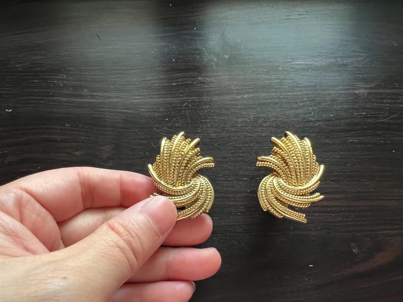 K&ouml;nnte beinhalten: Ein Paar goldfarbene Clip-Ohrringe. Jeder Ohrring hat ein strukturiertes, wirbelndes Design mit einer Ansammlung kleiner, vertikaler Linien. Die Ohrringe werden auf einer dunklen Holzoberfl&auml;che pr&auml;sentiert.