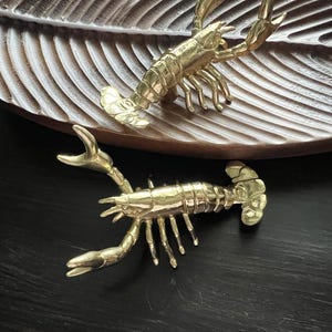 May include: Two brass lobster figurines rest on a wooden leaf-shaped tray. The lobsters are detailed with claws, legs, and segmented bodies. The tray has a dark brown finish and is set against a dark background.