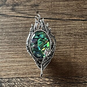 May include: An oval pendant with a silver-tone frame, featuring an iridescent abalone shell centre. The frame is embellished with small, dark stones and a feather-like design. It hangs from a thin black cord.