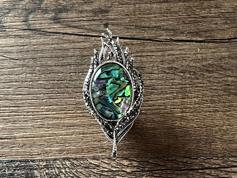 May include: An oval pendant with a silver-tone frame, featuring an iridescent abalone shell centre. The frame is embellished with small, dark stones and a feather-like design. It hangs from a thin black cord.
