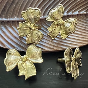 May include: Four gold-colored bow-shaped cabinet pulls. The pulls are made of metal and have a detailed, textured design. The bows are arranged on a wooden leaf-shaped tray and a black surface.