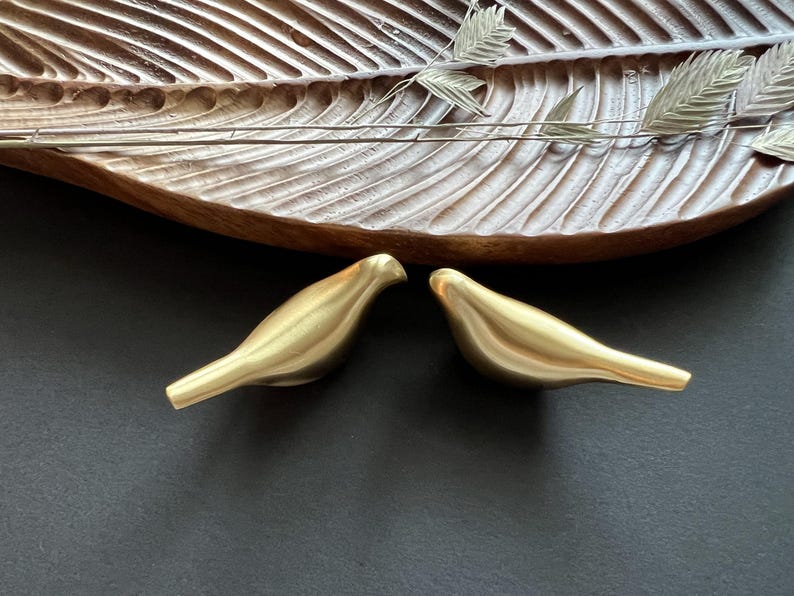 Pu&ograve; includere: Due figurine di uccelli in metallo dorato che si fronteggiano. Gli uccelli sono stilizzati e hanno una superficie liscia e arrotondata.