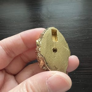 May include: Gold-colored metal decorative knob with a detailed design. The knob has a round shape and a hole in the center.