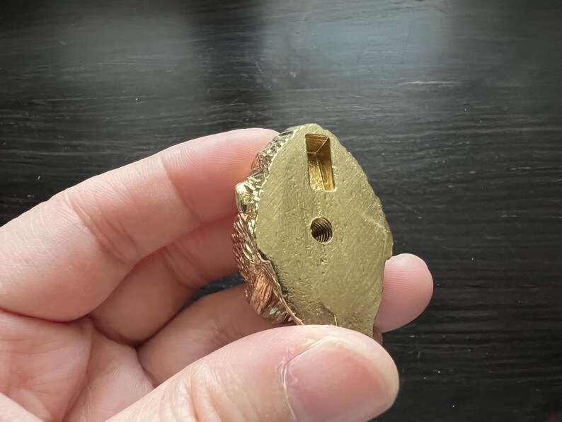 May include: Gold-colored metal decorative knob with a detailed design. The knob has a round shape and a hole in the center.