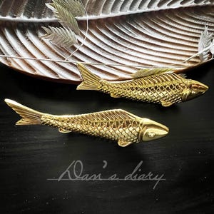 May include: Two gold-coloured fish-shaped decorative drawer pulls. The pulls are on a dark brown surface.