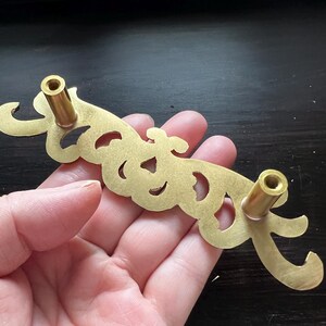 May include: A gold-toned metal drawer pull with a decorative, ornate design. The pull has two round knobs on either end.