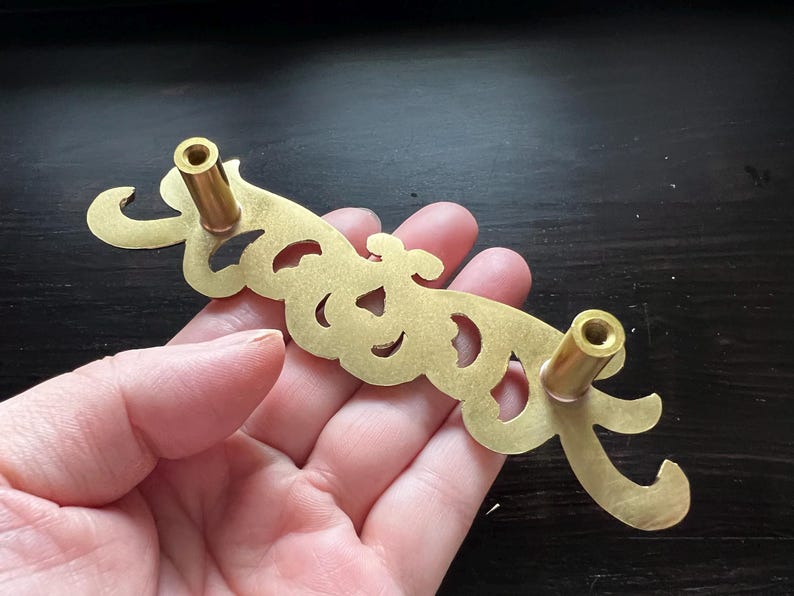 May include: A gold-toned metal drawer pull with a decorative, ornate design. The pull has two round knobs on either end.