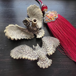 K&ouml;nnte beinhalten: Zwei Eulenfiguren aus Bronze mit ausgebreiteten Fl&uuml;geln. Eine hat eine zylindrische &Ouml;ffnung. Eine rote Quaste mit dekorativem floralem Akzent ist ebenfalls vorhanden. Die Artikel werden auf einer dunklen Holzoberfl&auml;che ausgestellt.