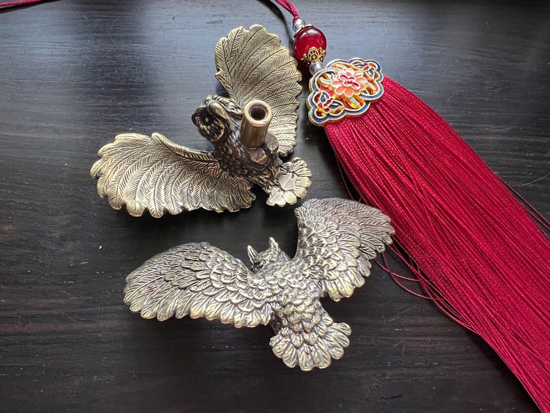 K&ouml;nnte beinhalten: Zwei Eulenfiguren aus Bronze mit ausgebreiteten Fl&uuml;geln. Eine hat eine zylindrische &Ouml;ffnung. Eine rote Quaste mit dekorativem floralem Akzent ist ebenfalls vorhanden. Die Artikel werden auf einer dunklen Holzoberfl&auml;che ausgestellt.