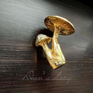 May include: Gold mushroom-shaped drawer pull. The pull is made of two mushrooms, one on top of the other. The top mushroom is larger and has a more rounded cap. The bottom mushroom is smaller and has a more pointed cap.