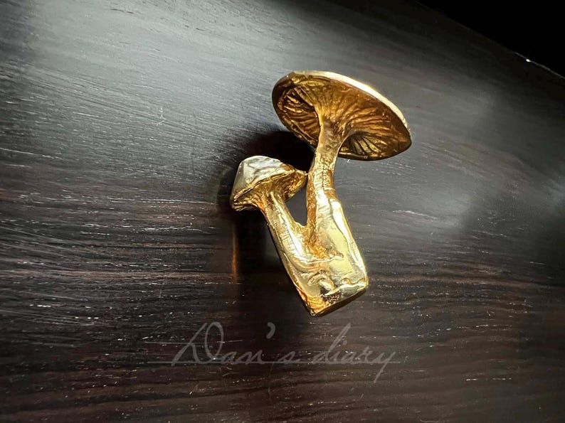 May include: Gold mushroom-shaped drawer pull. The pull is made of two mushrooms, one on top of the other. The top mushroom is larger and has a more rounded cap. The bottom mushroom is smaller and has a more pointed cap.