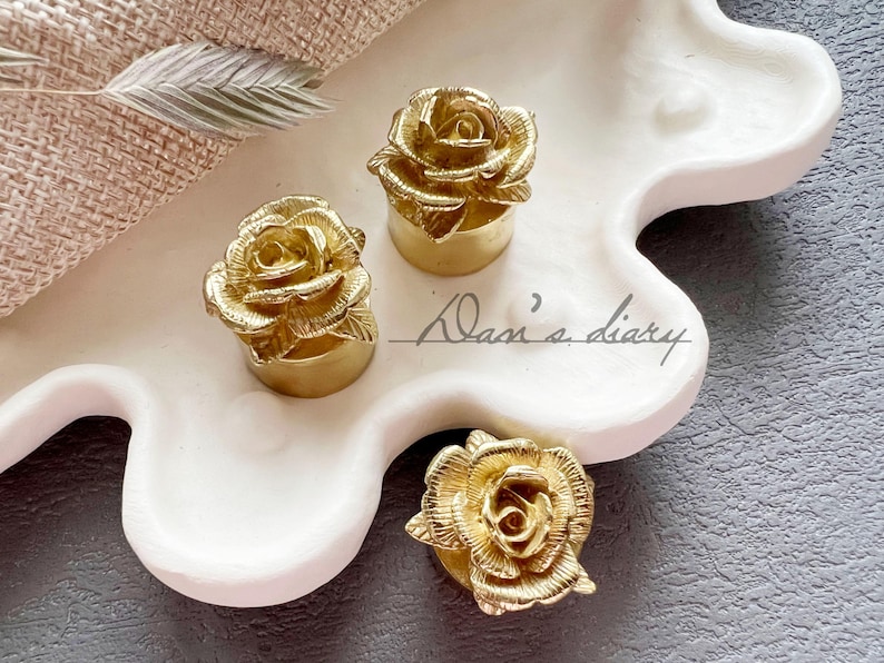 May include: Three gold-colored rose-shaped bottle stoppers on a white ceramic tray. The tray has a wavy edge.