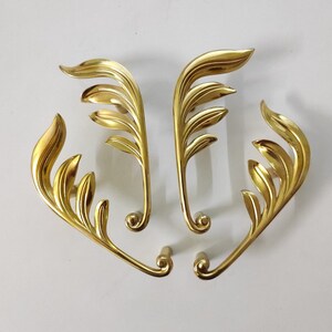 May include: Four gold-toned metal drawer pulls with a leaf design. Each pull has a curved shape and a decorative leaf pattern.