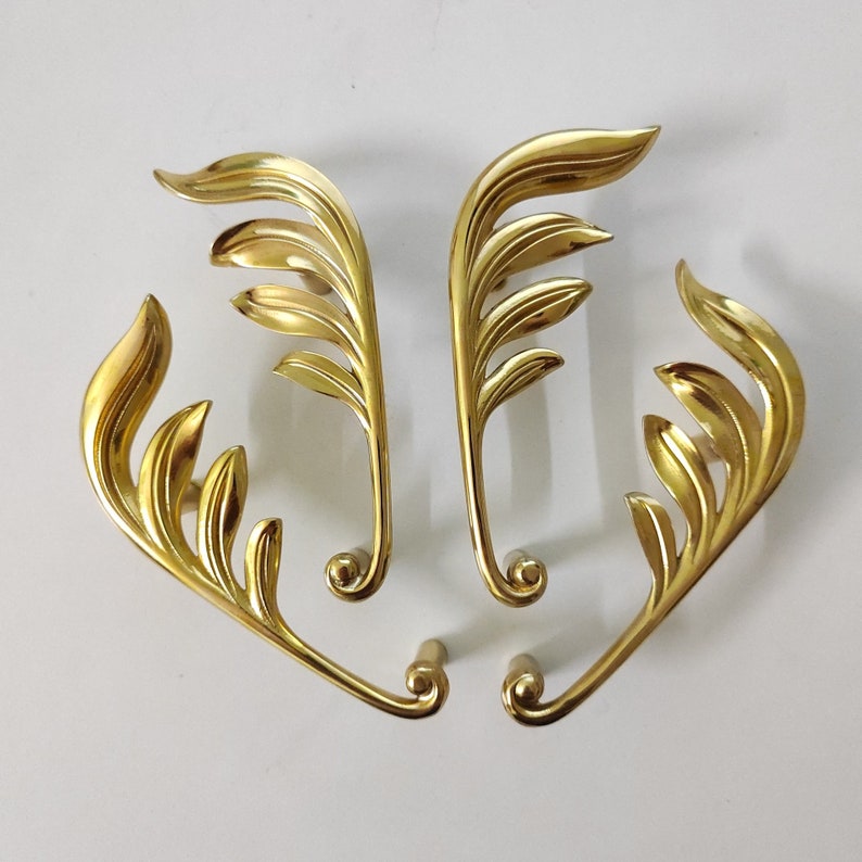 May include: Four gold-toned metal drawer pulls with a leaf design. Each pull has a curved shape and a decorative leaf pattern.
