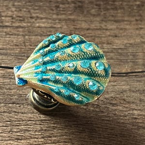 May include: A decorative seashell-shaped knob with a teal and gold finish, embellished with teal rhinestones. The knob is mounted on a bronze base, ideal for adding a coastal touch to furniture or decor.