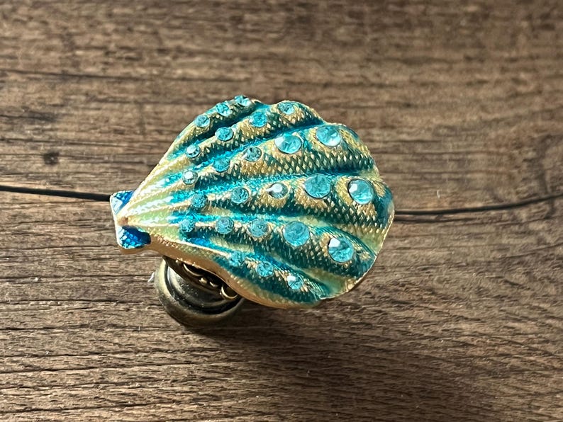 May include: A decorative seashell-shaped knob with a teal and gold finish, embellished with teal rhinestones. The knob is mounted on a bronze base, ideal for adding a coastal touch to furniture or decor.