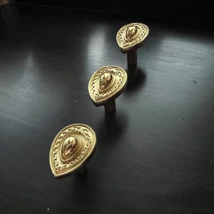 May include: Three gold-toned decorative cabinet knobs with a teardrop shape and intricate detailing. The knobs have a raised center and a textured, ornate design.