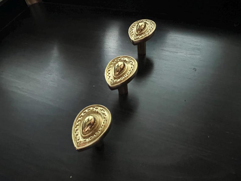May include: Three gold-toned decorative cabinet knobs with a teardrop shape and intricate detailing. The knobs have a raised center and a textured, ornate design.