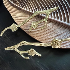 Puede incluir: Dos tiradores de armario de metal dorado en forma de ramas estilizadas con detalles de flores.