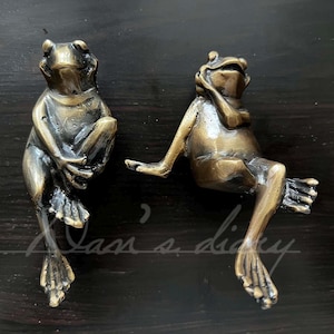 May include: Two brass frog figurines, one sitting with its legs crossed and the other sitting with its legs extended. Both frogs have a realistic design and are made of a shiny brass material.