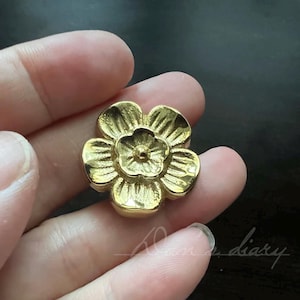 Könnte beinhalten: Ein goldfarbener, metallischer, blumenförmiger Dekorknopf mit erhabener Mitte.