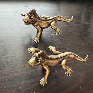 May include: Two brass lizard-shaped cabinet pulls. The lizards are in a crawling position with detailed scales and claws. The pulls are a warm gold color and are set against a dark wood surface.