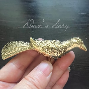 May include: A gold-colored bird-shaped decorative object. The bird has detailed feather and wing patterns, with a slightly open beak. The object is held in a hand against a dark background. The text "Dan's diary" is written in cursive.