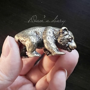 May include: A small, detailed bronze bear figurine. The bear is in a walking pose, with textured fur and realistic features. The figurine is held in a hand, with the text "Dan's diary" in the background.