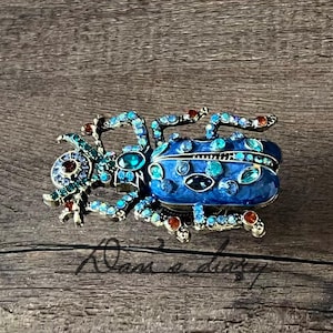 May include: A decorative blue jeweled beetle figurine with turquoise and red accents. The beetle is set on a wooden surface. The figurine has a hinged top and is likely a trinket box.
