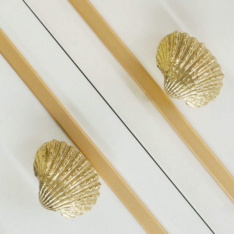 May include: Two gold seashell drawer pulls on a white surface. The seashells are detailed and have a textured finish.