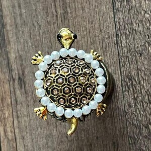 Puede incluir: Broche dorada con forma de tortuga y un diseño de caparazón geométrico detallado. La concha está adornada con pequeños cristales brillantes y rodeada por una fila de perlas blancas. La cabeza y las patas de la tortuga también son doradas.