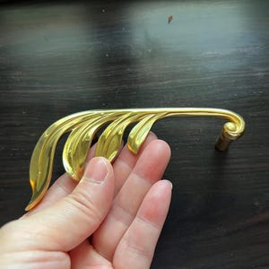 May include: Gold-toned metal cabinet pull with a leaf design. The pull is shaped like a branch with multiple leaves.