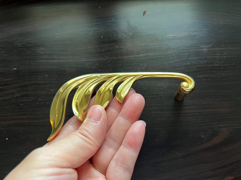 May include: Gold-toned metal cabinet pull with a leaf design. The pull is shaped like a branch with multiple leaves.