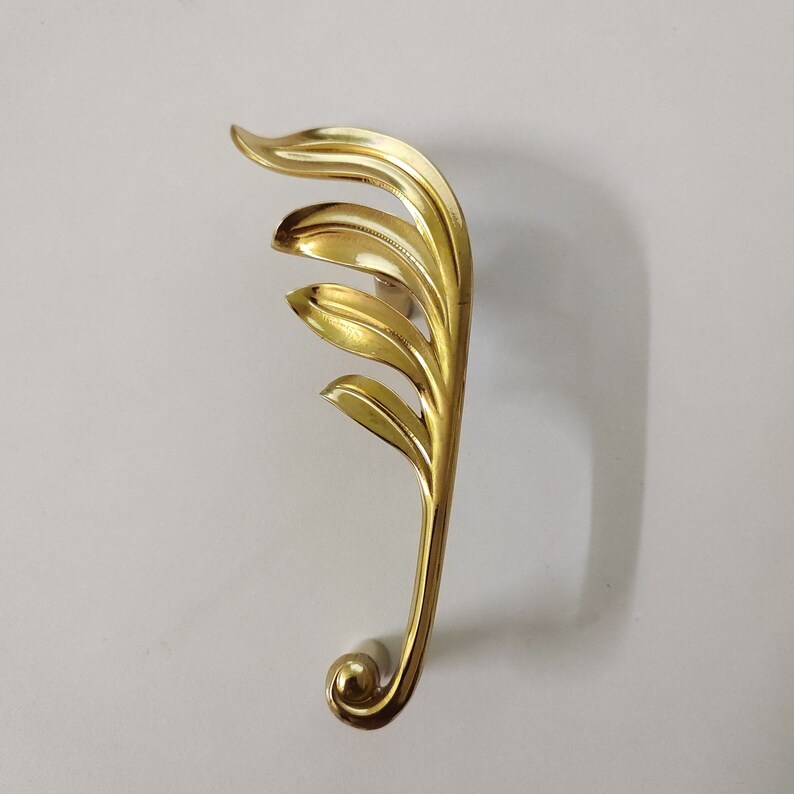 May include: A gold-toned door handle with a leaf design. The handle is curved and has a rounded end.