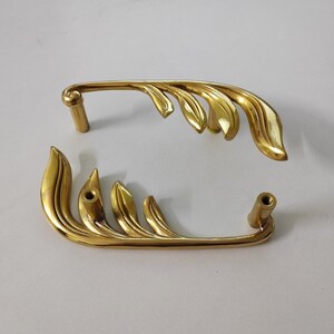 May include: Two gold-toned metal cabinet door pulls with a leaf design. The pulls are in the shape of a stylized leaf with a rounded end.