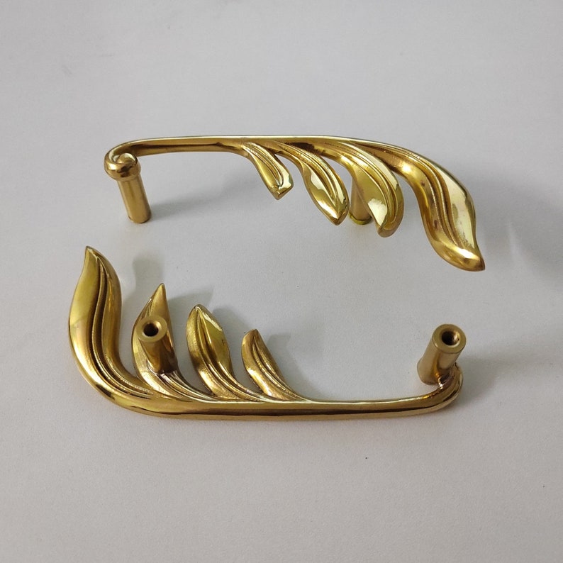 May include: Two gold-toned metal cabinet door pulls with a leaf design. The pulls are in the shape of a stylized leaf with a rounded end.