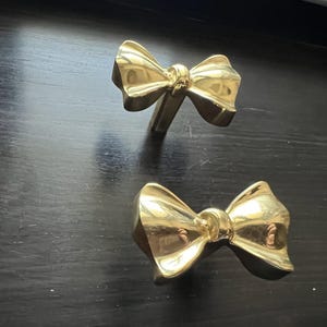 May include: Two gold-colored bow-shaped drawer pulls. The pulls are made of metal and have a shiny finish.