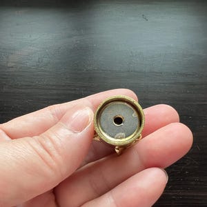 May include: A gold-colored metal base with a round top and a small hole in the center. The base has a decorative design around the edge.