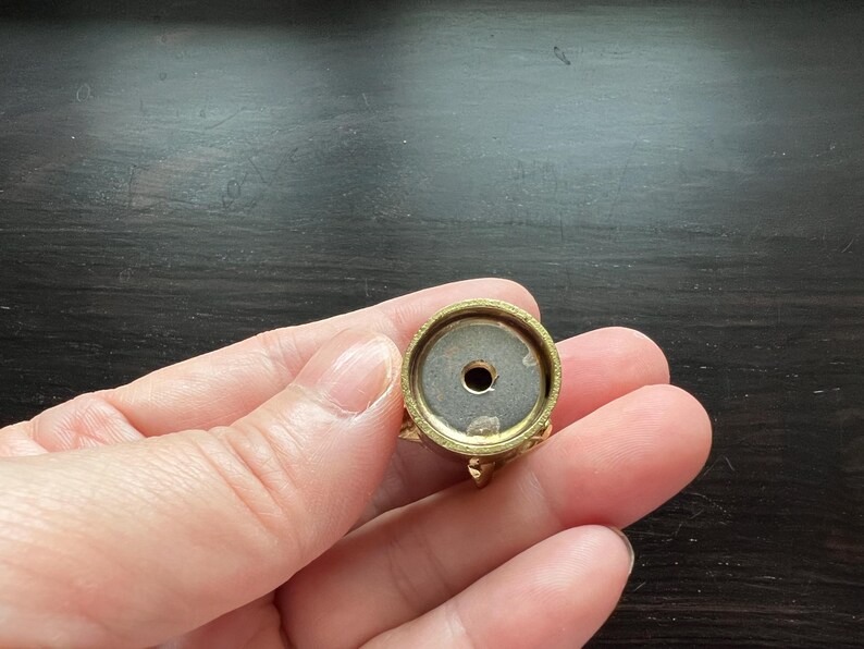 May include: A gold-colored metal base with a round top and a small hole in the center. The base has a decorative design around the edge.