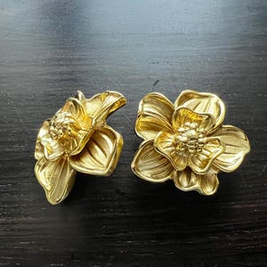 May include: A pair of gold-tone flower-shaped earrings. The earrings are made of metal and have a detailed design.