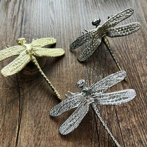 May include: Three decorative dragonfly figurines in gold, silver, and bronze tones. The dragonflies have detailed wing patterns and slender bodies. They are arranged on a wooden surface.